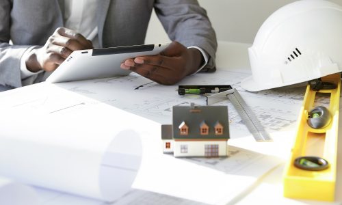 Cropped shot of dark-skinned contractor holding touch pad, entering data while working on new housing project, sitting at desk with drawings, scale model house, blueprint rolls, ruler and helmets